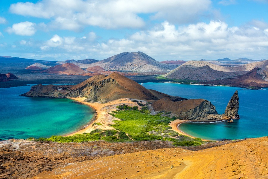 Una formación volcánica con dos bahías se extiende entre aguas turquesas y colinas áridas bajo un cielo azul con nubes blancas.