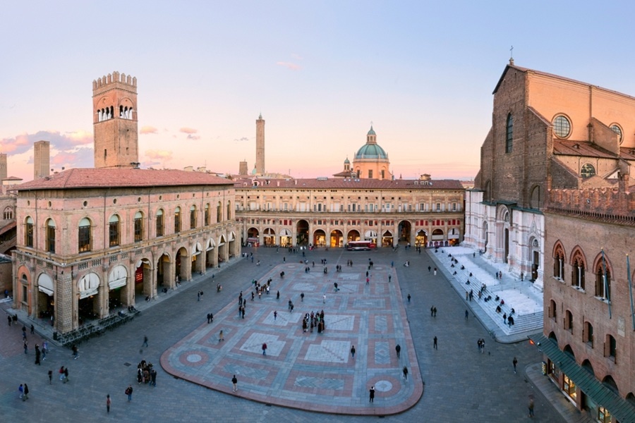Varias personas caminan y descansan por la gran Piazza Maggiore de Bolonia, rodeada de edificios históricos de terracota, la basílica de San Petronio y torres medievales bajo el atardecer.