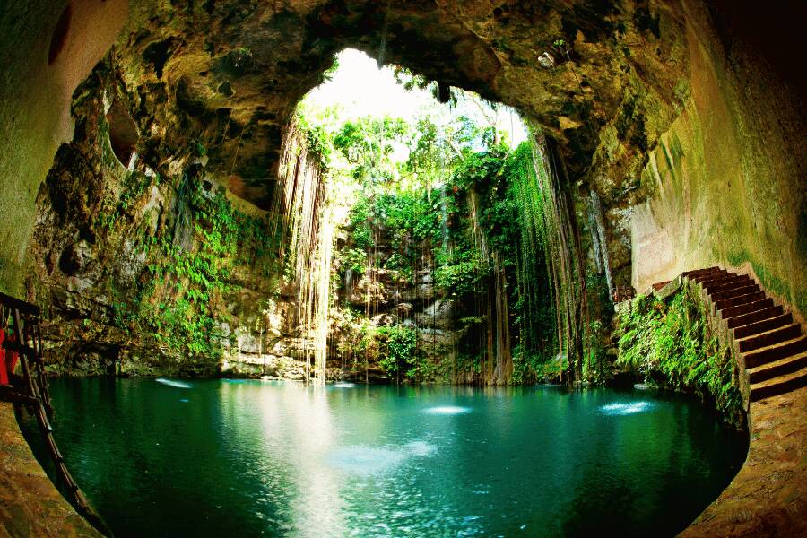 La luz del sol se filtra por una abertura rocosa sobre un cenote subterráneo con escaleras de piedra en el lateral.