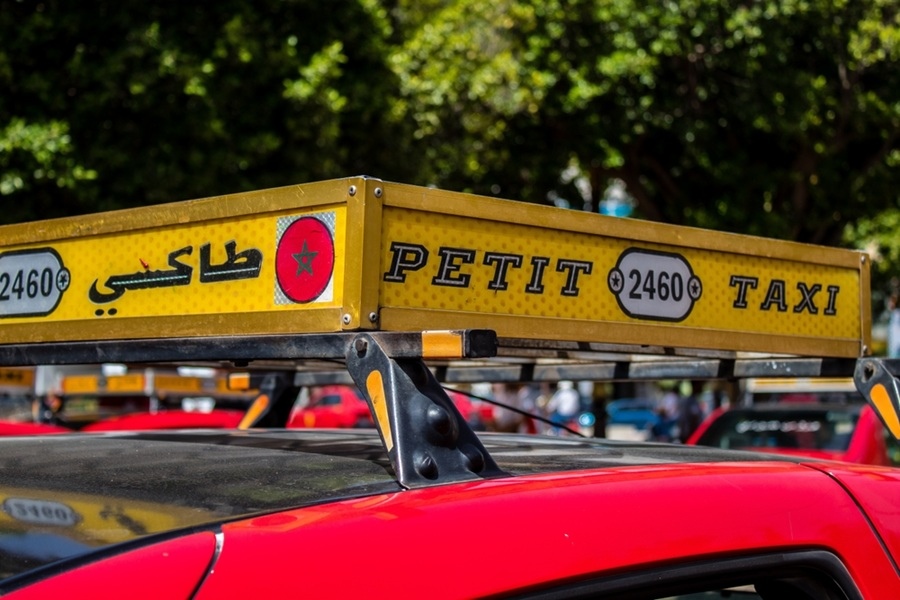 Un letrero amarillo sobre el techo de un taxi rojo exhibe los textos «طاكسي», «PETIT TAXI» y el número «2460», con vegetación de fondo.