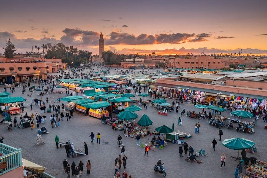 Numerosos puestos de mercado y transeúntes se extienden por una gran plaza abierta al atardecer, con edificios de tonos cálidos y un alminar destacando al fondo.