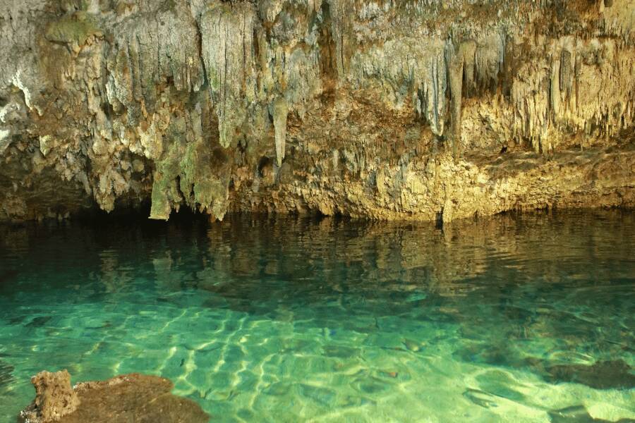 El agua cristalina de un estanque se calma bajo el techo rocoso de una cueva con estalactitas colgantes.