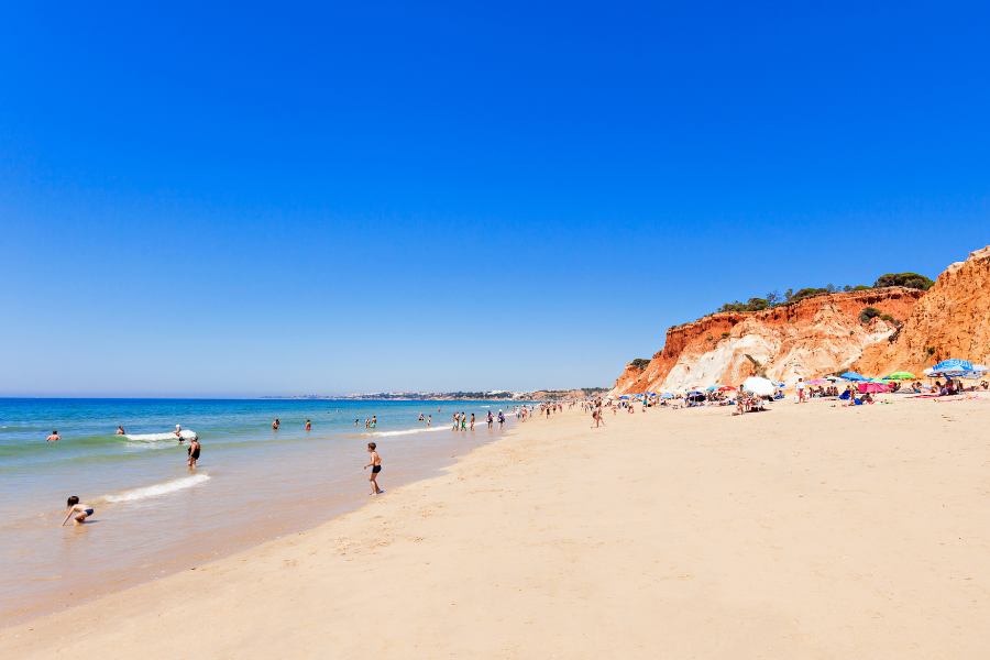 Playa con turistas bañandose y de fondo los majestuosos acantilados de arcilla roja y ocre junto a la orilla del océano Atlántico en Praia da Falesia en el Algarve de Portugal.