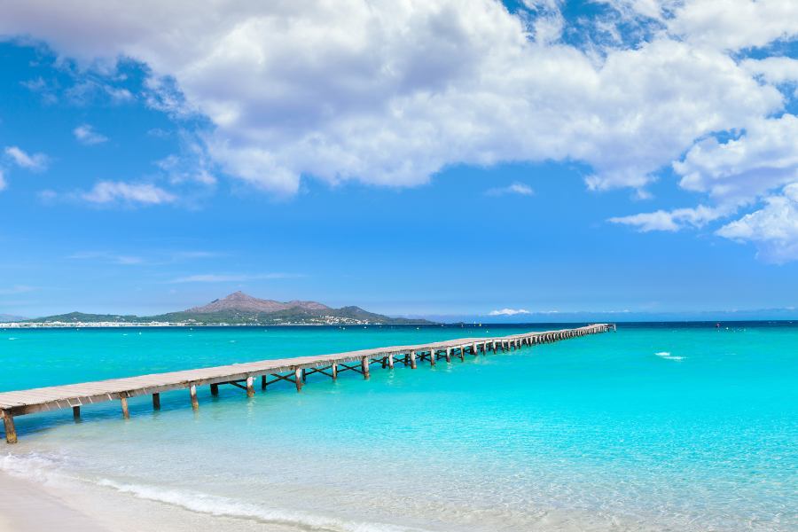 Extenso arenal y aguas cristalinas someras en Playa de Muro Mallorca con sus tradicionales pantalanes de madera adentrándose en el mar.