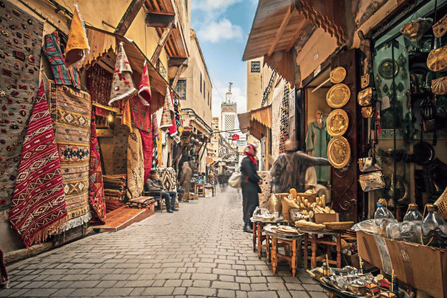 Alfombras y artesanías tradicionales cuelgan a los lados de un concurrido zoco de piedra mientras varias personas caminan por el pasaje de la medina. Fez, marruecos.