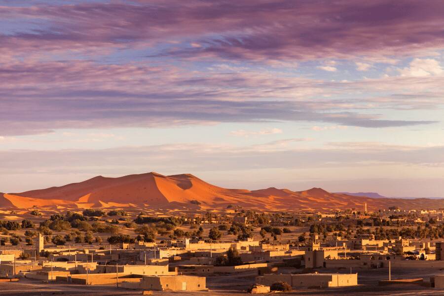 Unas doradas dunas de arena se extienden tras las edificaciones de barro de un pueblo del desierto bajo el cielo del atardecer. Merzouga.