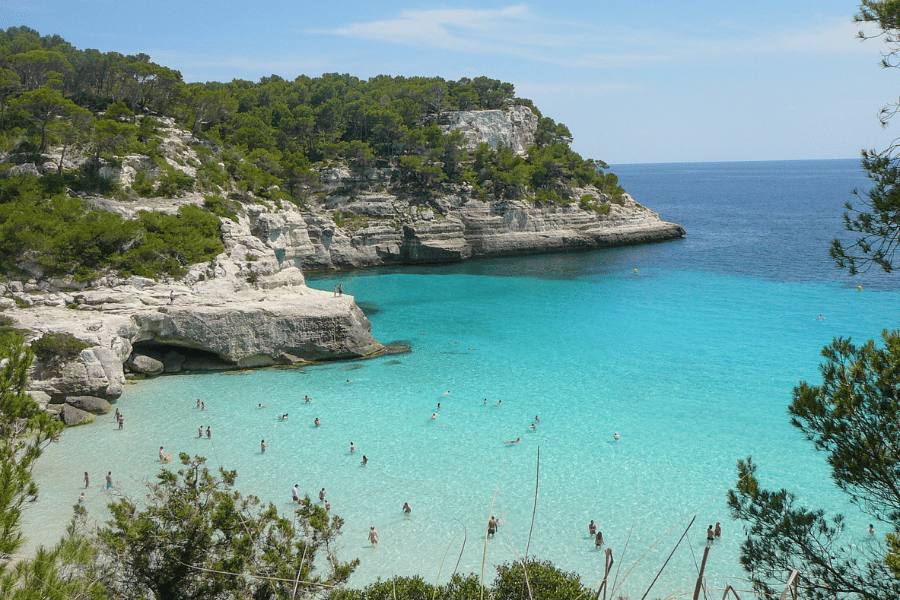 Cala de agua turquesa con bañistas, enmarcada por acantilados cubiertos de vegetación, en la isla de Menorca, España, en un día despejado.