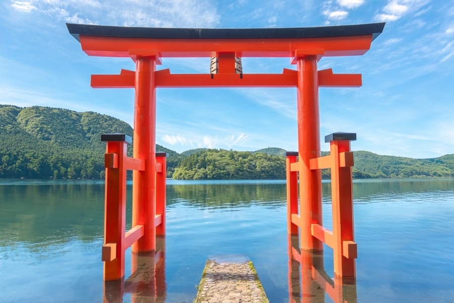 Una gran puerta torii de color rojo bermellón se alza sobre el agua de un lago tranquilo, rodeada de frondosas colinas verdes bajo un cielo despejado.