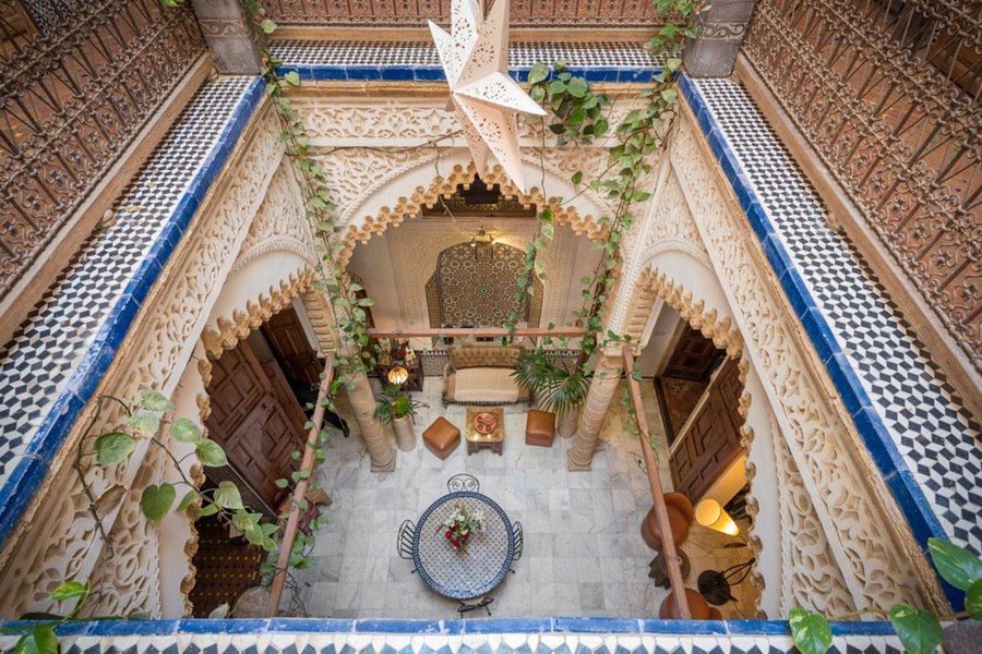 Una lámpara en forma de estrella cuelga sobre un patio interior tradicional, decorado con arcos tallados, plantas trepadoras, azulejos geométricos y zonas de descanso amuebladas.