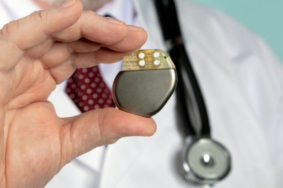 Un marcapasos cardíaco metálico es sostenido entre los dedos de un médico, quien viste bata blanca, corbata roja y estetoscopio, sugiriendo una explicación o revisión de este dispositivo médico.