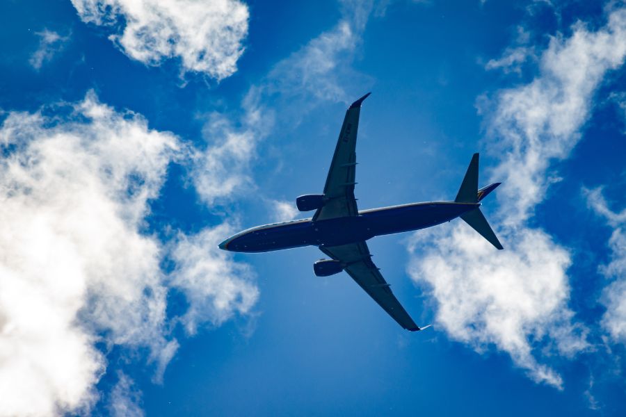 Un avión comercial vuela directamente sobre el observador, con sus alas y motores detallados contra un cielo azul intenso salpicado de nubes blancas algodonosas.
