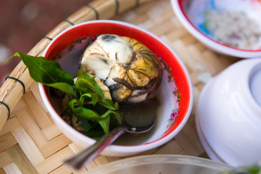 Un huevo balut cocido y abierto reposa sumergido en caldo junto a hojas verdes dentro de un cuenco pequeño de borde rojo colocado sobre una bandeja de mimbre.
