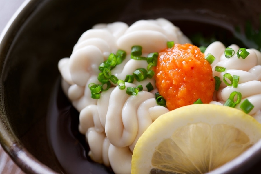 Un porción de shirako blanco reposa servida en un cuenco oscuro junto a una rodaja de limón, cebolleta picada y una cucharada de salsa ankimoko.