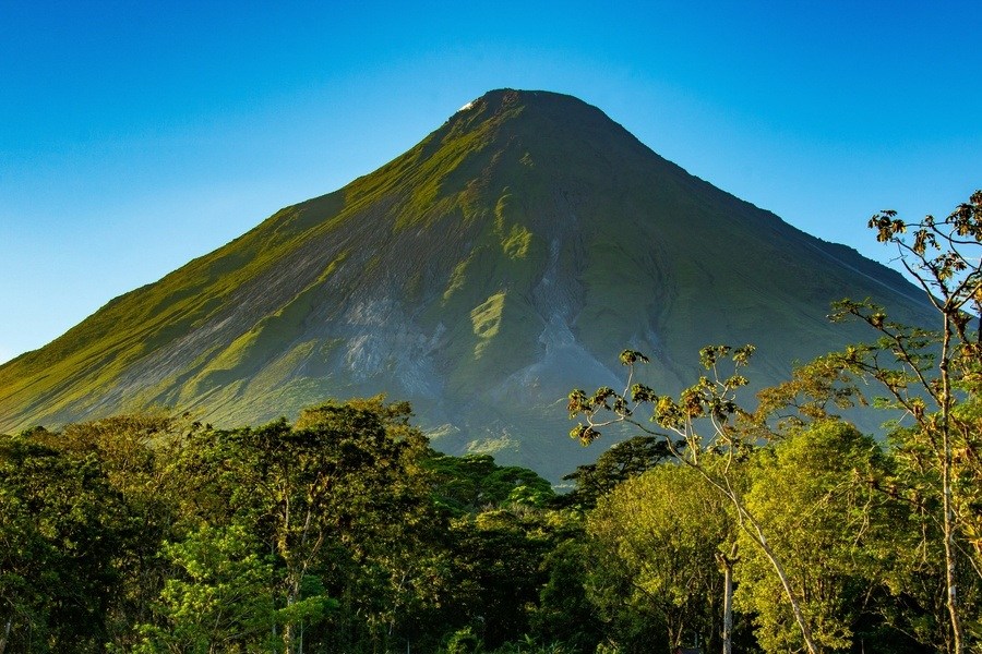 Un gran volcán cónico de laderas verdes se erige majestuoso hacia un cielo despejado, alzándose por encima de un denso bosque tropical de frondosos árboles bajo la luz del sol.