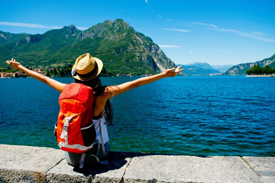 Mujer con sombrero de paja y mochila roja se sienta de espaldas en un muro de piedra contemplando un gran lago azul rodeado de montañas bajo un cielo despejado, disfrutando de la tranquilidad de viajar con insulina.