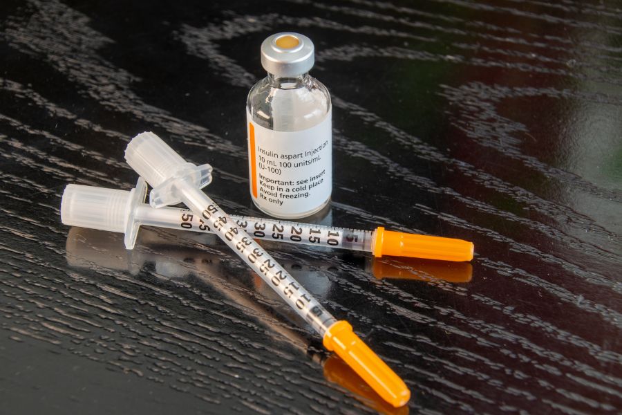 Dos plumas de insulina con capuchones blancos y agujas finas acopladas reposan sobre una superficie lisa de color rosa claro, mostrando marcas numéricas impresas que indican 60, 120 y 180 para planificar un viaje seguro con diabetes.