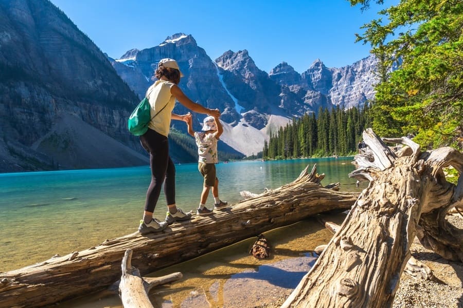 Madre e hijo balanceándose en un tronco erosionado por el vibrante lago de morrena turquesa, rodeado por los majestuosos picos de las rocas canadienses en un soleado día de verano