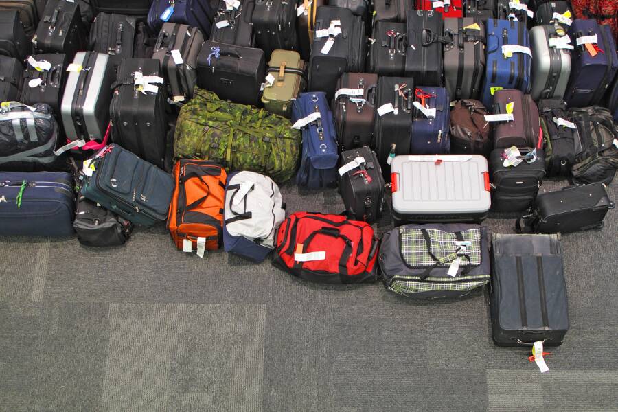 Faqs heymondo Numerosas maletas, mochilas y bolsas de viaje de diferentes colores y tamaños aparecen agrupadas y ordenadas en el suelo de moqueta gris de la terminal de un aeropuerto.