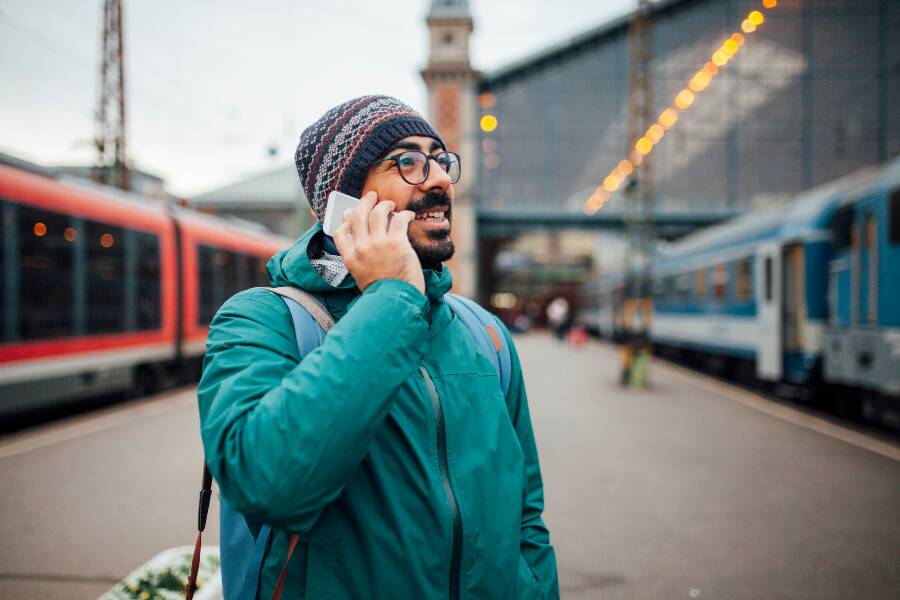 Un hombre con gafas, gorro de lana y chaqueta verde habla por su teléfono móvil mientras sonríe, situado en el andén exterior de una estación ferroviaria entre dos trenes de pasajeros.