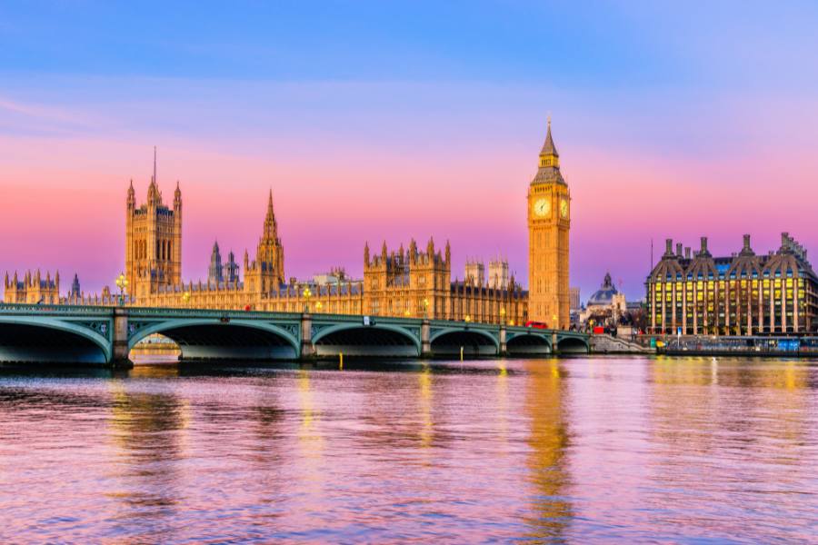 El palacio del parlamento con su emblemática torre del reloj se sitúa junto a un río bajo un cielo con tonos rosados y morados al atardecer.