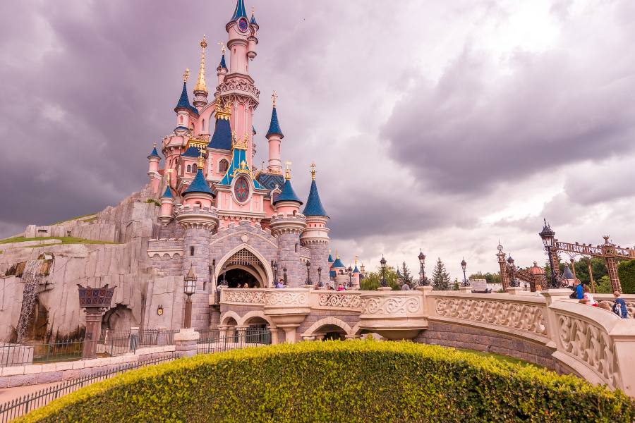 Un gran castillo de cuento de hadas con tejados azules y agujas doradas se alza detrás de setos verdes bajo un cielo nublado.