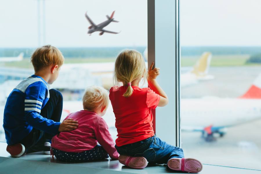 Tres niños sentados junto a un gran ventanal de aeropuerto observan de espaldas un avión comercial despegando en la pista exterior bajo un cielo claro.
