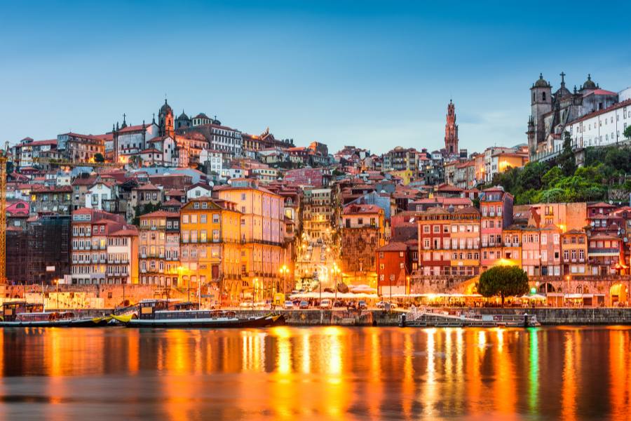 Edificios antiguos de colores iluminados en una colina se reflejan sobre la superficie del agua de un río durante el atardecer.