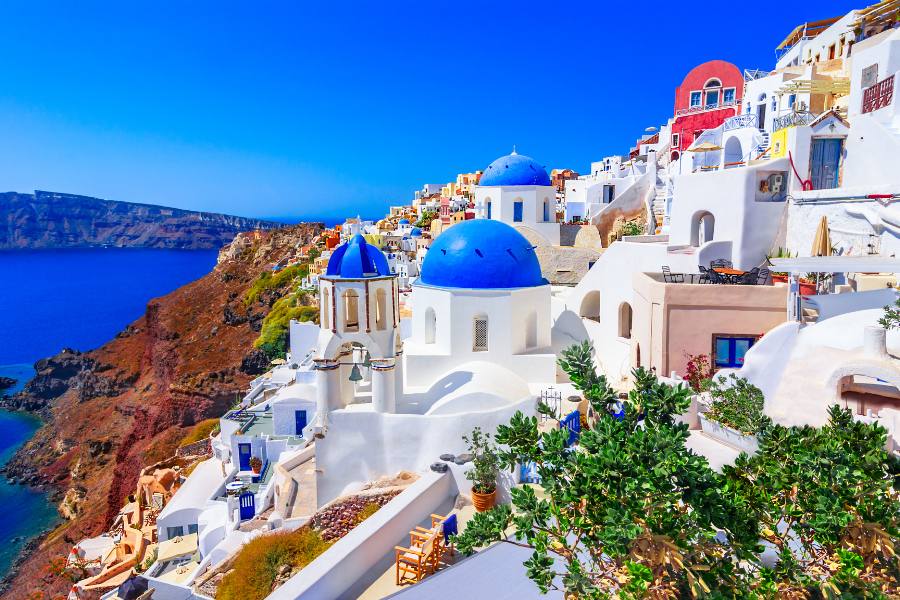 Edificios blancos con cúpulas azules se extienden sobre un acantilado escarpado junto al mar azul brillante bajo un cielo totalmente despejado.