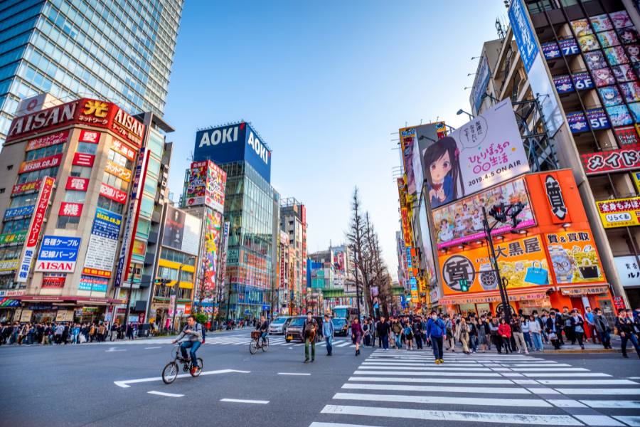 Personas cruzan un paso de peatones y un ciclista avanza en una avenida urbana rodeada de edificios altos con carteles publicitarios comerciales. Texto visible: "AOKI", "AISAN", "2019.4.5 ON AIR".