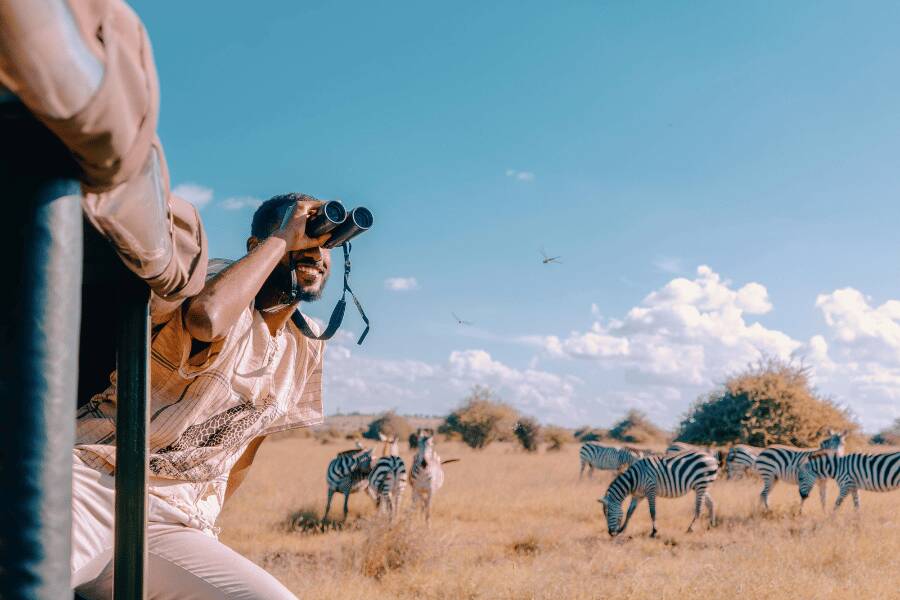 Un turista observa zebras con prismáticos en medio de la sabana africana bajo un cielo despejado con nubes dispersas.