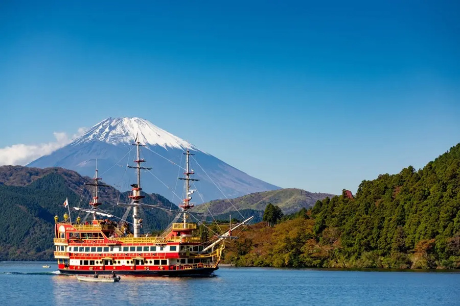 Un barco turístico de estilo pirata navega por un lago tranquilo, con el imponente monte Fuji nevado al fondo y frondosas colinas arboladas a los lados.