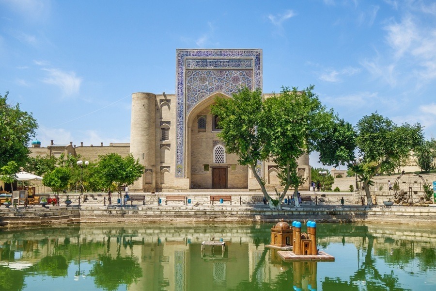Fachada monumental de la madraza Nadir Divan-begi reflejada en el estanque central del complejo Lyabi-Khauz, rodeado de árboles verdes bajo un cielo claro en Bujará, Uzbekistán.