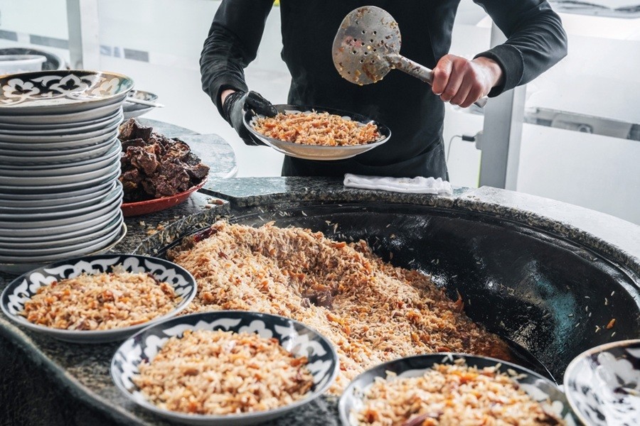 Un cocinero sirve plov tradicional uzbeko con una espumadera desde un enorme caldero hacia varios platos decorados con motivos locales.