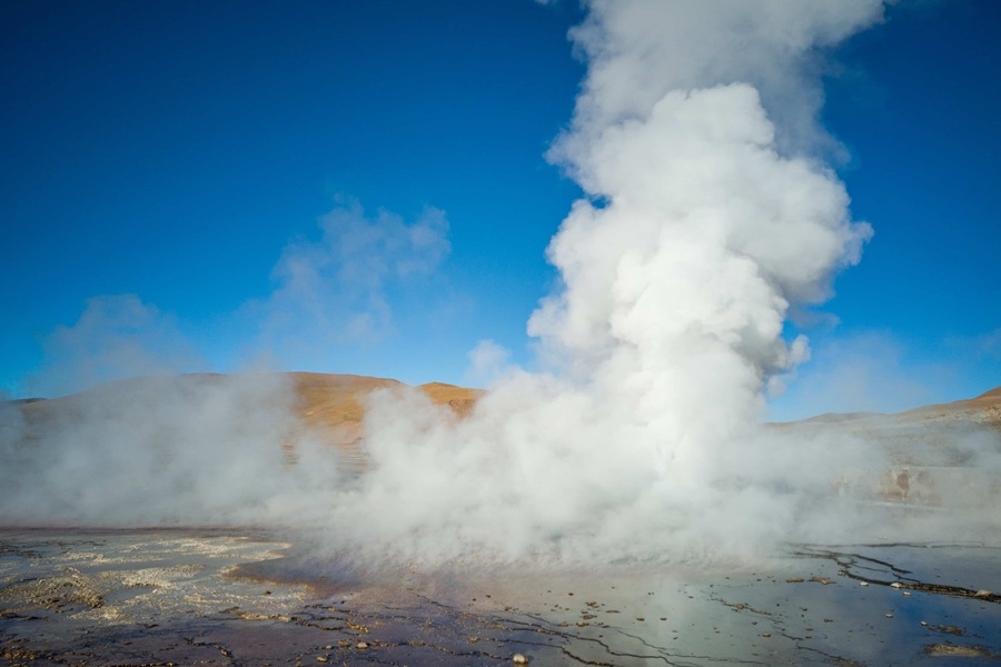 Una densa columna de vapor blanco brota violentamente del suelo en un campo geotérmico, rodeada de terreno árido y colinas bajo un cielo azul despejado.