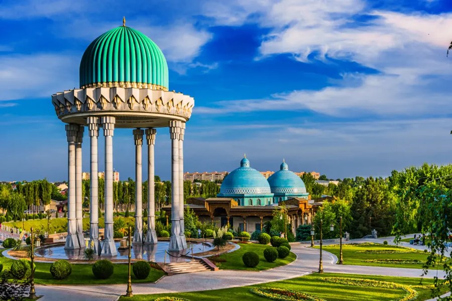 Glorieta con cúpula verde sostenida por columnas blancas sobre un estanque, junto a un edificio de cúpulas azules en un parque ajardinado en Tashkent, Uzbekistán.