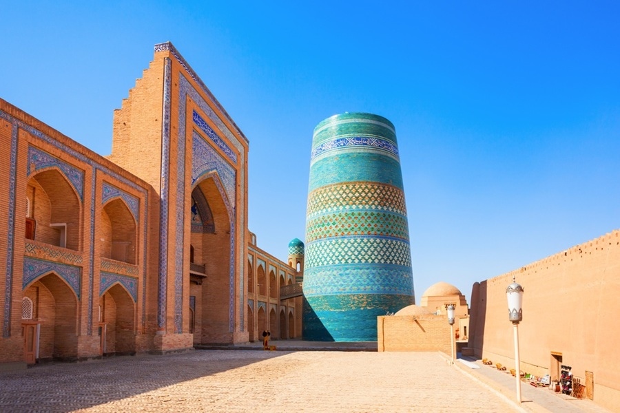 El minarete icónico de Kalta Minor, recubierto de azulejos turquesas y verdes, se alza junto a una madraza de ladrillo y murallas de barro en la ciudad amurallada de Itchan Kala, Jiva.