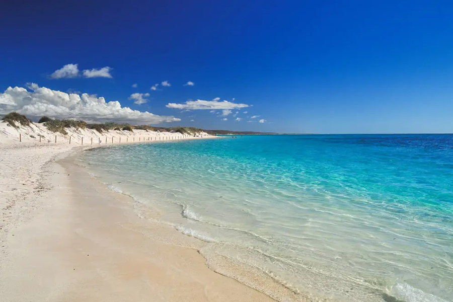 Aguas turquesas y cristalinas bañan suavemente una orilla de arena blanca junto a dunas costeras, bajo un cielo despejado de color azul Intenso.