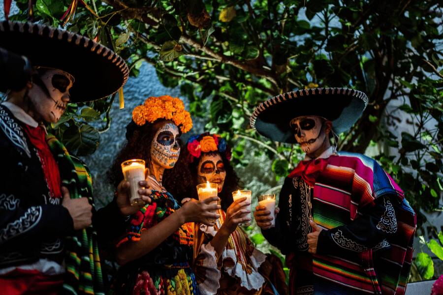 Un grupo de personas caracterizadas de Catrines sostiene velas encendidas durante la noche rodeado de vegetación.