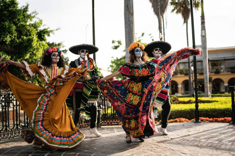 Bailarines caracterizados como Catrines y Catrinas danzan con vestidos coloridos tradicionales en una plaza pública al aire libre.