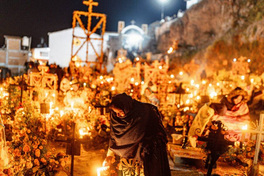 Personas envueltas en mantas velan a sus difuntos junto a tumbas iluminadas con velas y cruces en un cementerio nocturno.