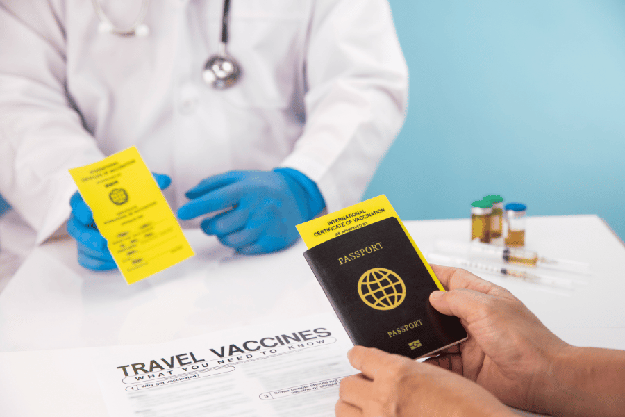 Un sanitario con guantes azules entrega un pasaporte y un certificado de vacunación amarillo a un paciente sobre un escritorio con viales médicos.