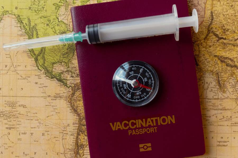 Jeringuilla médica y brújula de bolsillo reposan sobre un pasaporte de vacunación rojo colocado sobre un mapa terráqueo antiguo.