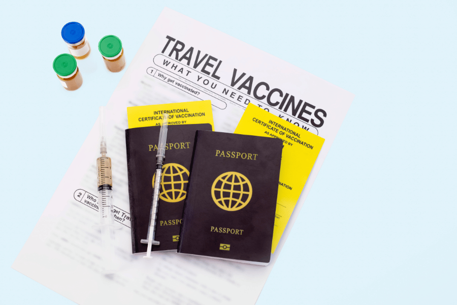 Dos pasaportes negros, un certificado de vacunación amarillo, una jeringuilla y varios viales de vacunas reposan sobre un folleto informativo en un fondo azul claro.