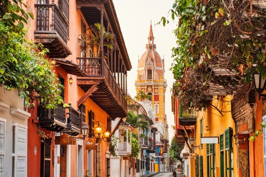 Una colorida torre catedralicia se alza al fondo de una estrecha calle colonial flanqueada por casas amarillas y naranjas con balcones de madera, farolas y enredaderas.