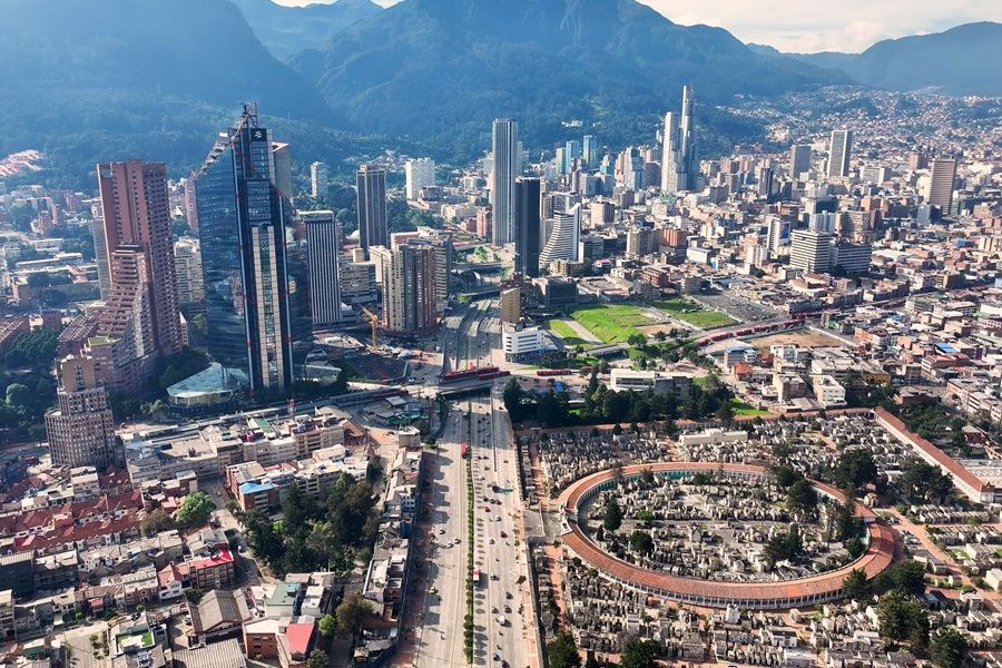Una amplia avenida con tráfico atraviesa un denso paisaje urbano con rascacielos y un cementerio circular, extendiéndose hacia montañas verdes bajo un cielo despejado.
