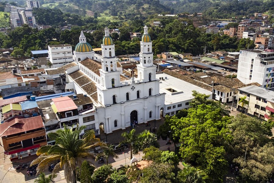 Una gran iglesia blanca con cúpulas de colores y altas torres destaca en el centro de un pueblo montañoso, rodeada de tejados diversos, frondosos árboles y colinas verdes