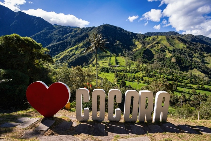 Un gran corazón rojo y letras volumétricas blancas con el texto «COCORA» se alzan sobre un mirador frente a colinas verdes, palmas de cera y montañas andinas bajo el cielo soleado.