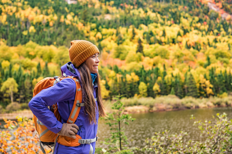 Una senderista con mochila y gorro camina de perfil en un bosque con árboles de hojas otoñales amarillas junto a un lago.
