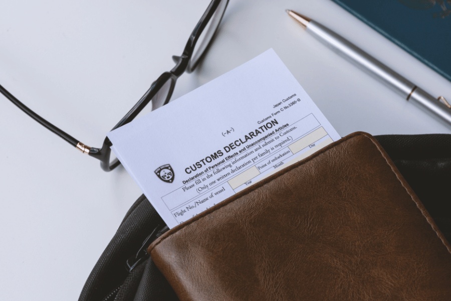 Gafas graduadas, un bolígrafo y una cartera de piel con un documento que contiene el texto «PASSENGER INFORMATION», dispuestos sobre una superficie blanca de trabajo.