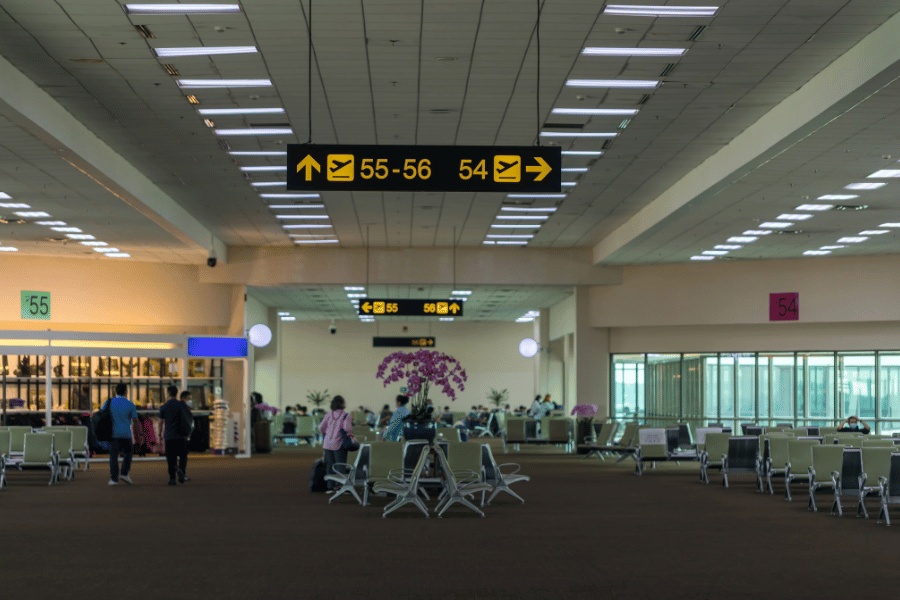 Pasillo de una terminal de aeropuerto iluminado con paneles digitales superiores que indican las puertas «50 - 59», mostradores al fondo y asientos de espera.